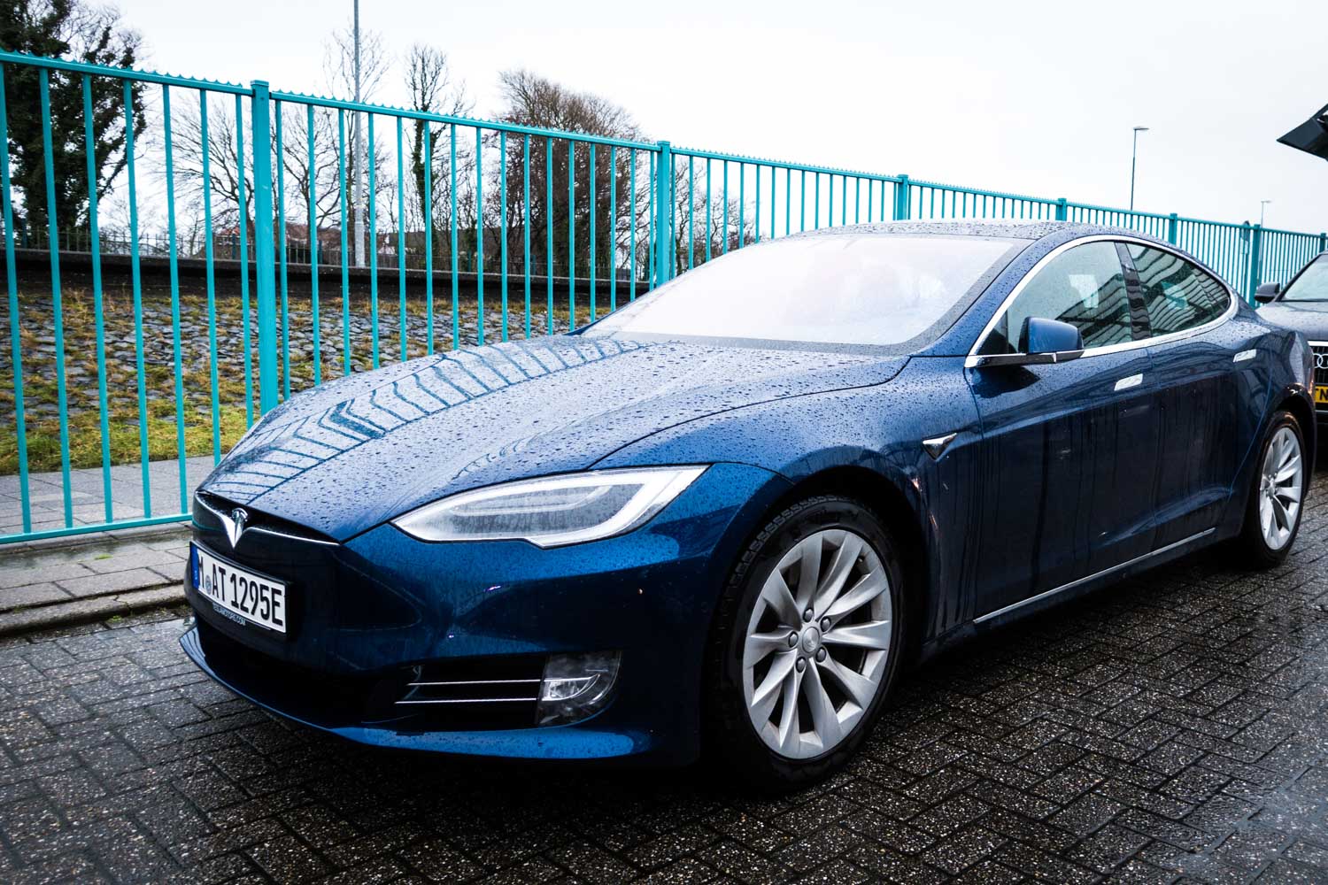 Blue Tesla Model S parked on wet pavement beside turquoise fence on a rainy day.
