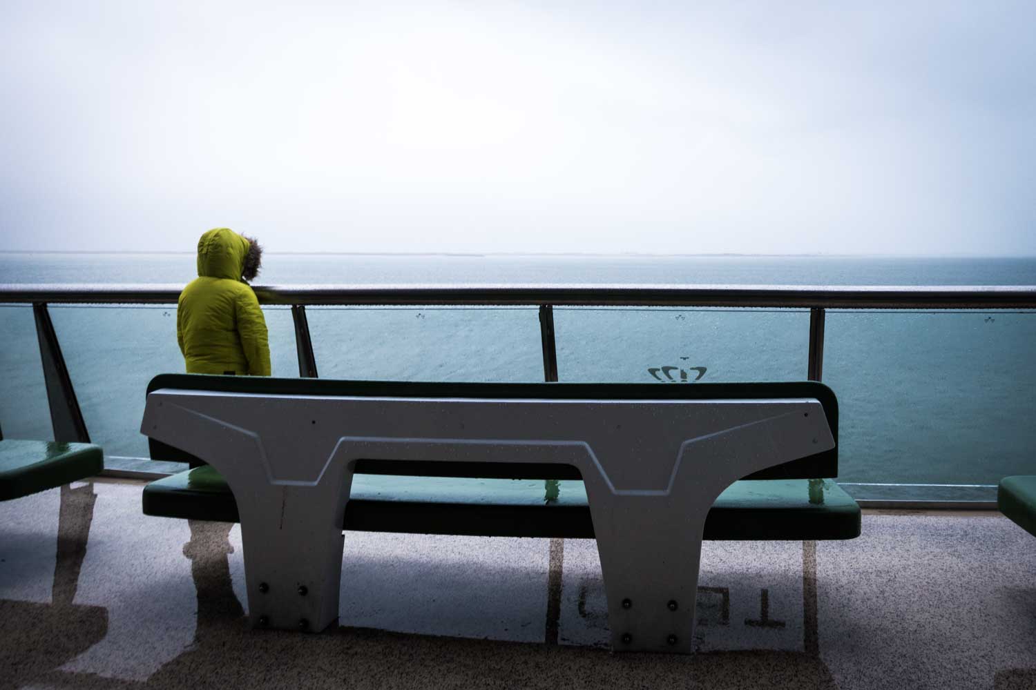 Person in yellow jacket on a ferry, gazing at a calm sea under a cloudy sky, with empty deck benches.