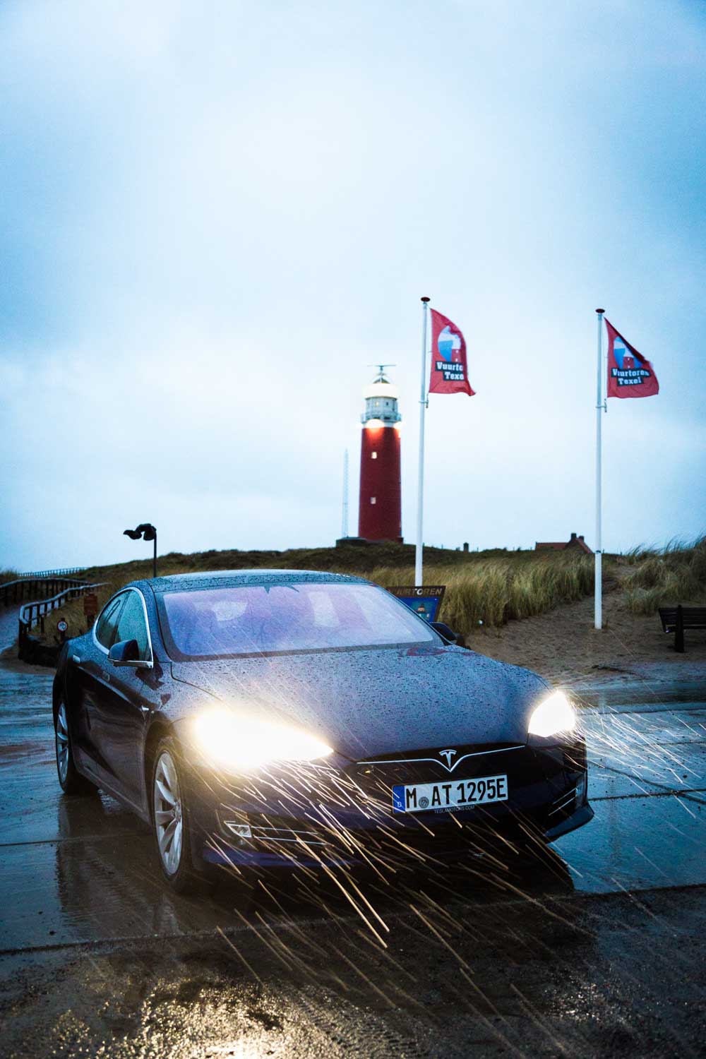 Tesla driving on a wet coastal road with a lighthouse and two flags in the background on a rainy day.