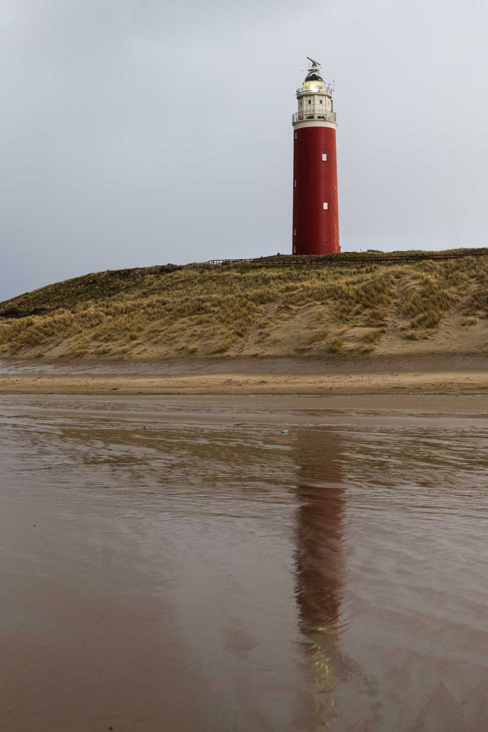 Red lighthouse on sandy hill reflects in calm beach water under cloudy sky.