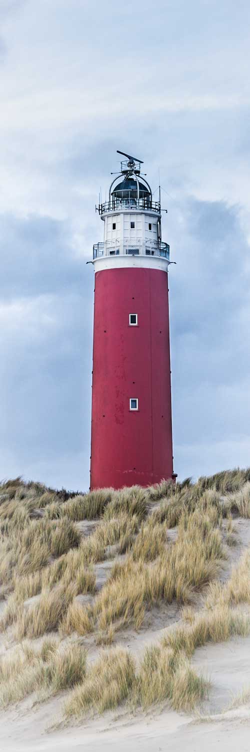 Red lighthouse tower above grassy dunes under a cloudy sky.