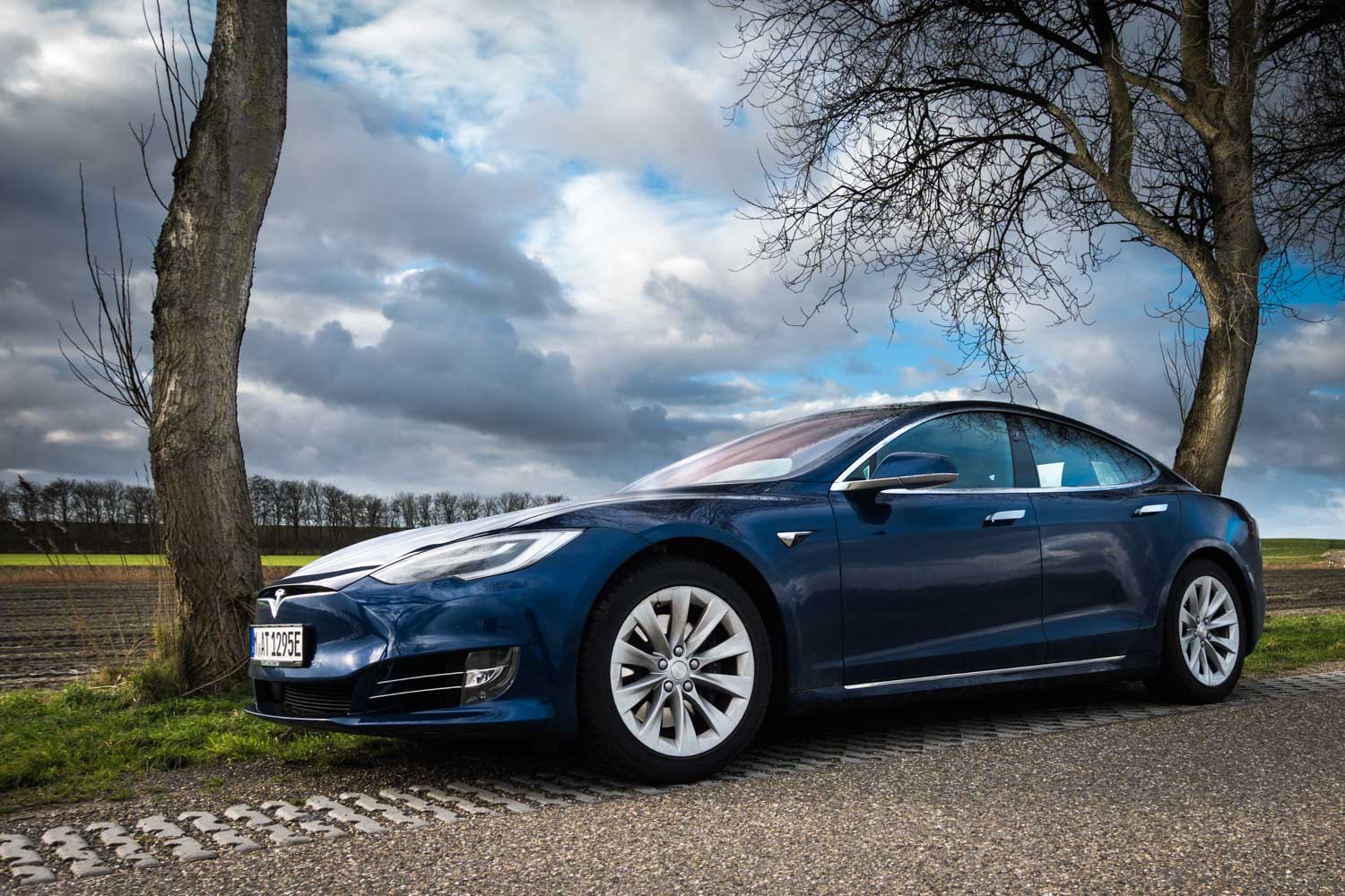 Blue Tesla car parked on rural road between trees under a cloudy sky.