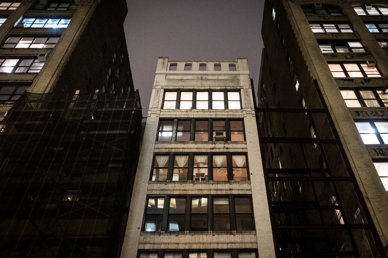 Tall city buildings at night with lit windows and dark sky above.