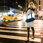 Woman in checkered coat crossing busy city street at night with yellow taxi speeding past.