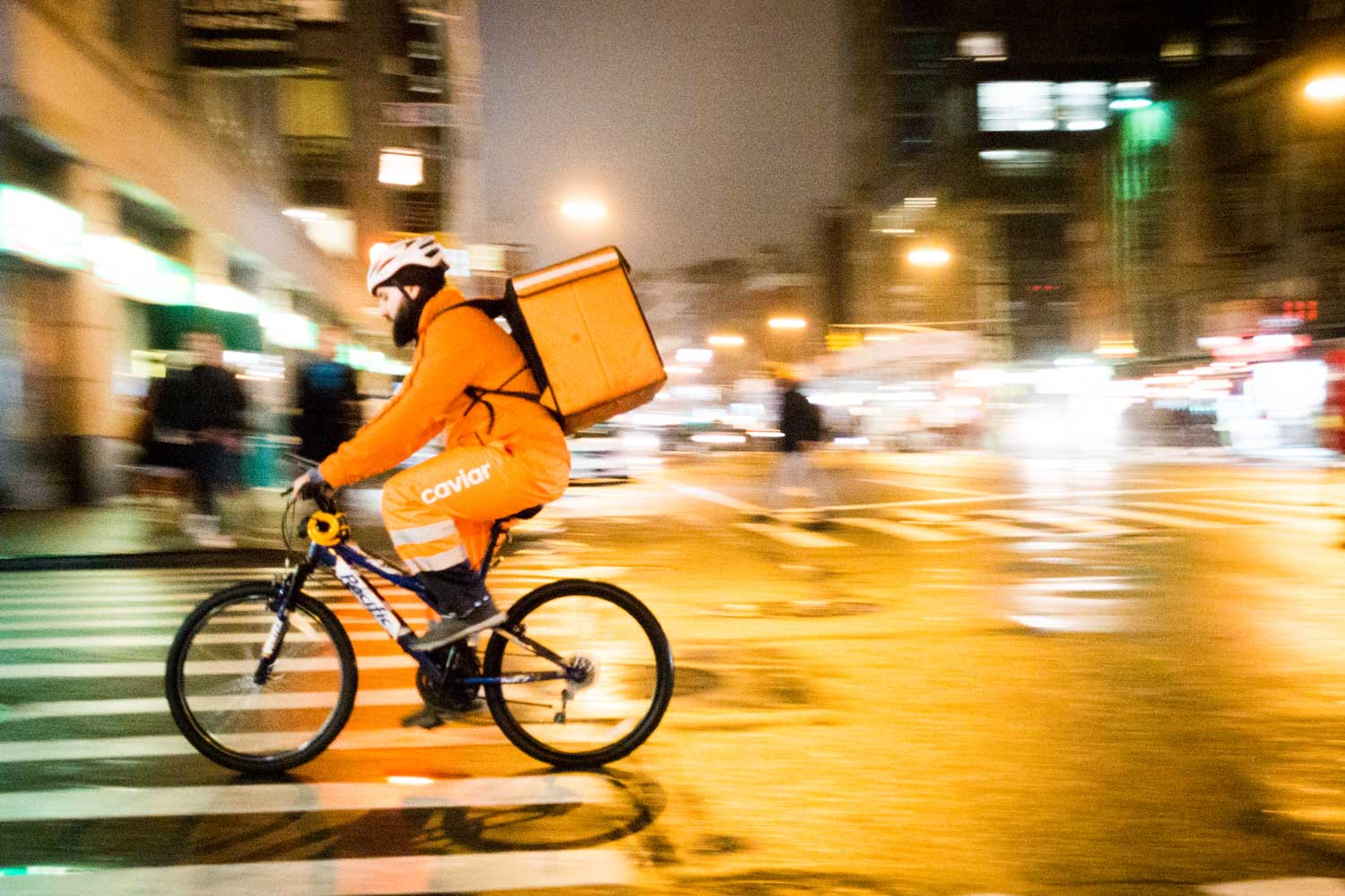 Delivery cyclist in orange jacket rides through city intersection at night.