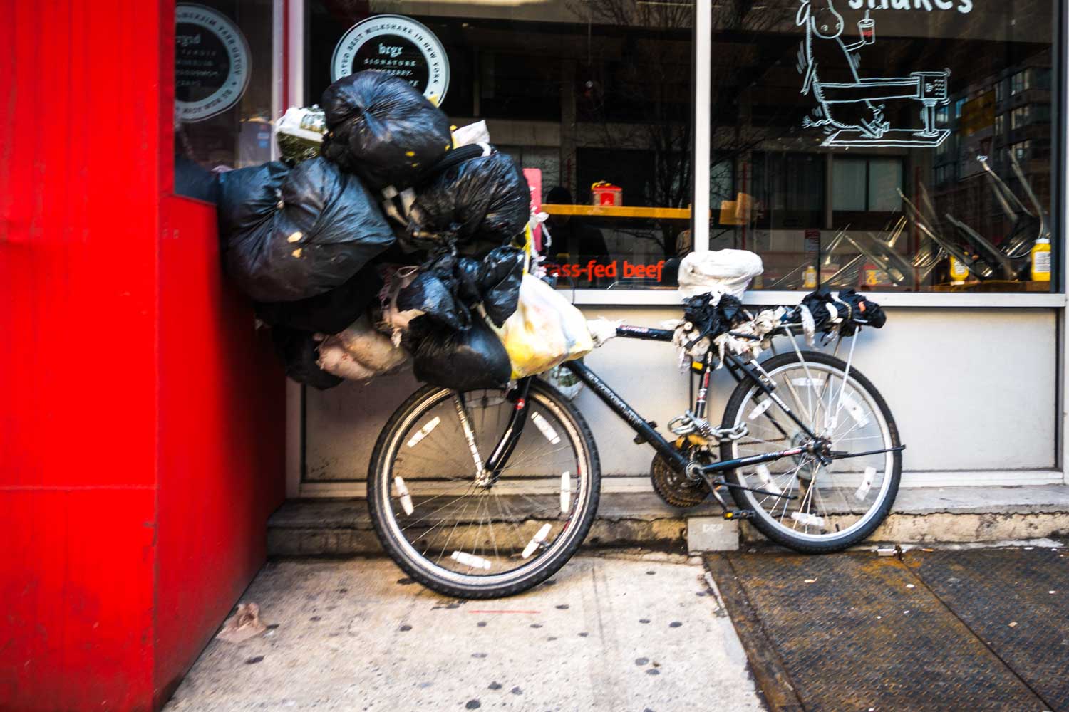 Bicycle laden with black garbage bags, parked against a red wall near a window with signage.