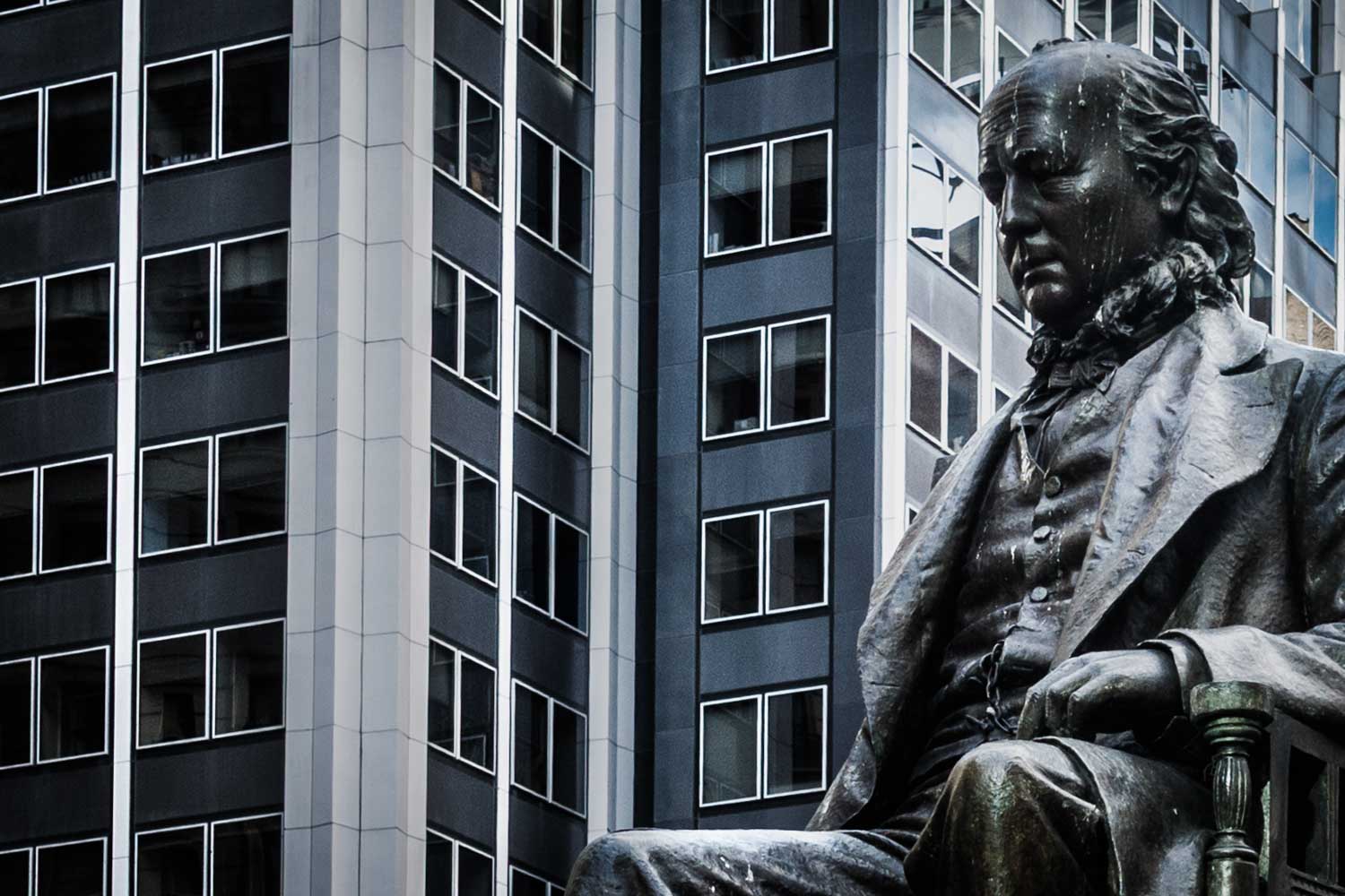 Statue of a seated man in front of modern high-rise buildings, showcasing urban contrast and historical significance.