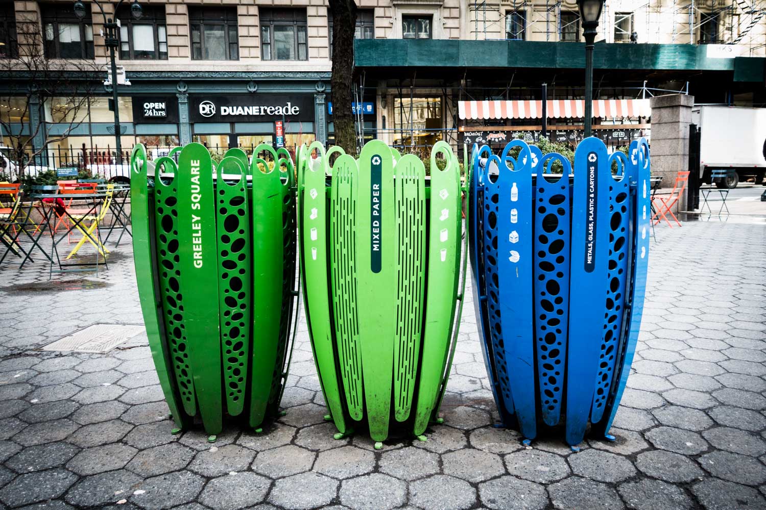 Green and blue recycling bins in Greeley Square, New York, with urban storefronts and cafe seating in the background.