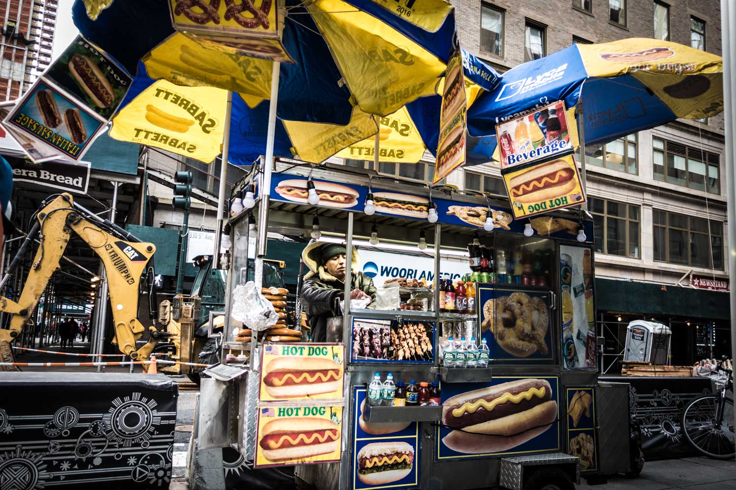 Street food cart in NYC selling hot dogs and pretzels under branded umbrellas with construction site background.