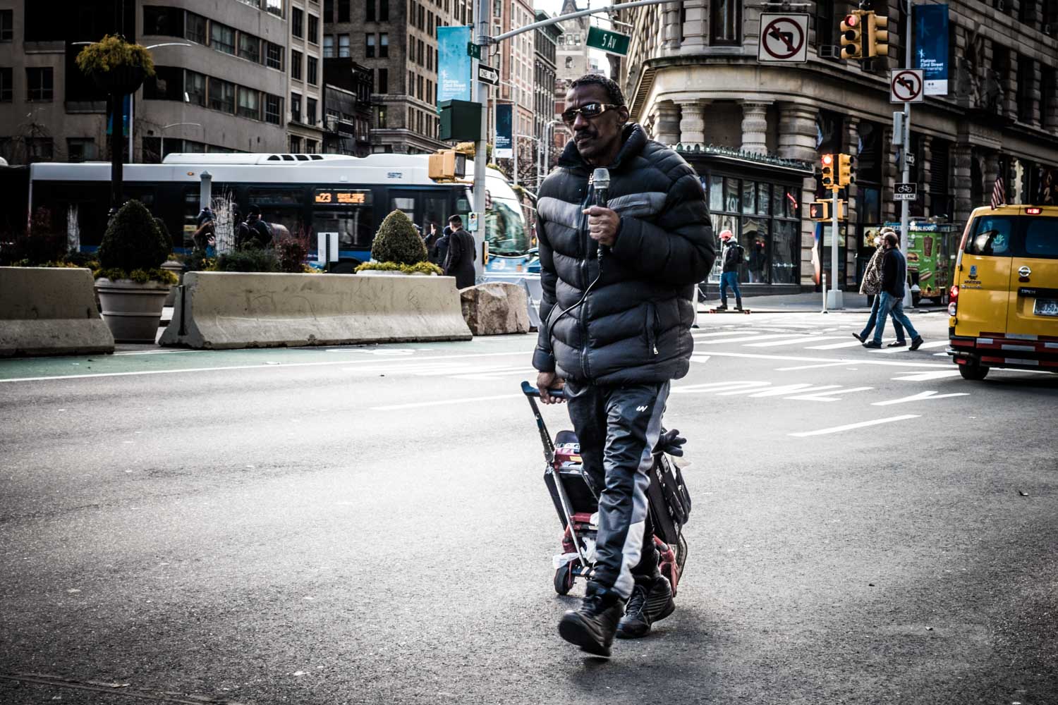 Man crossing a busy city street with a microphone, wearing a black jacket and pushing a cart, surrounded by urban signage.