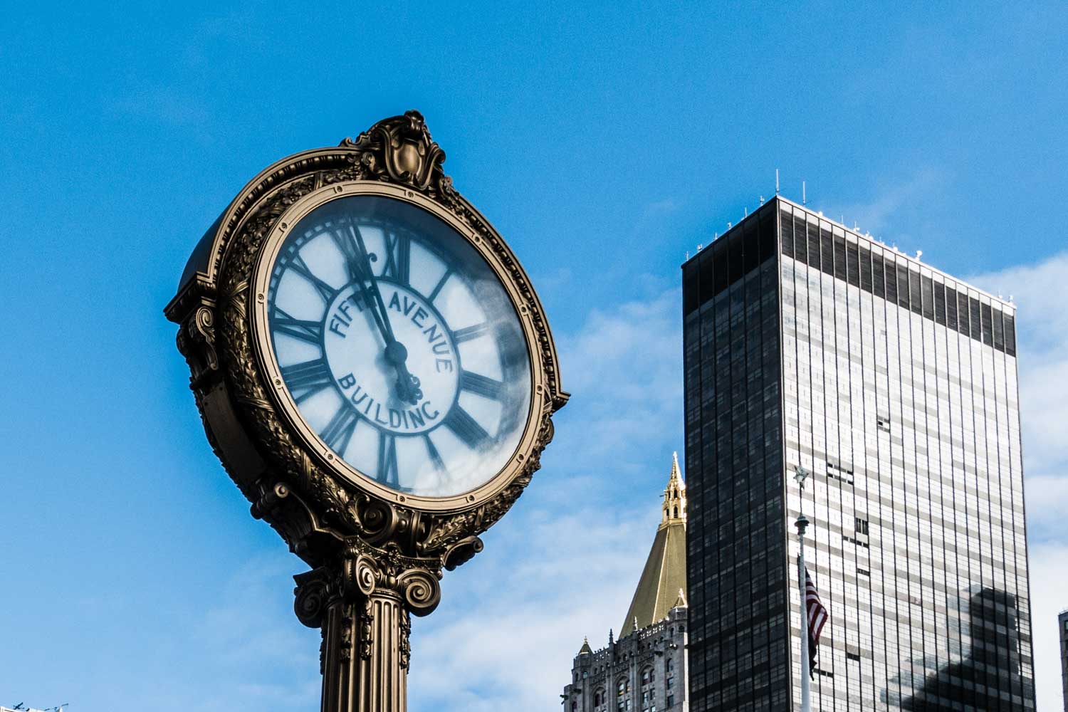 Ornate Fifth Avenue Building clock and skyscrapers against a clear blue sky.