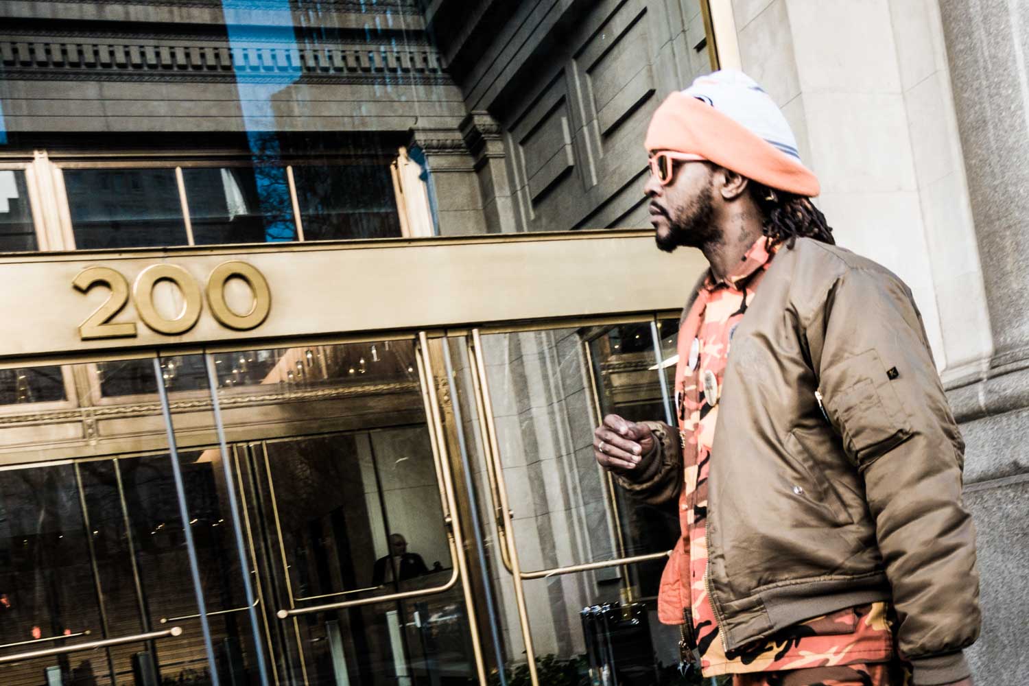 Man in orange beanie and bomber jacket walks past building entrance marked 200.