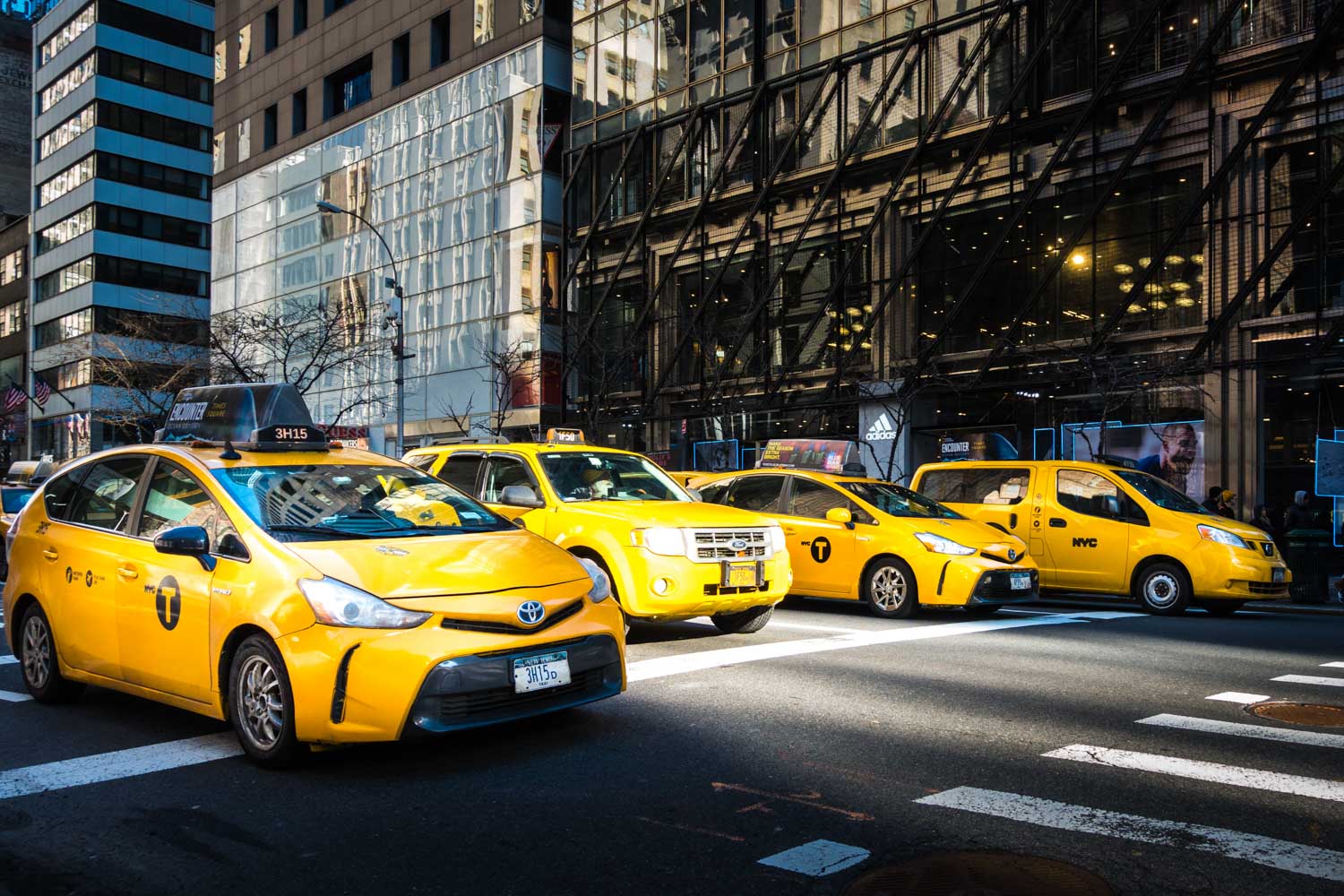Yellow taxis lined up on a busy New York City street with tall buildings in the background.
