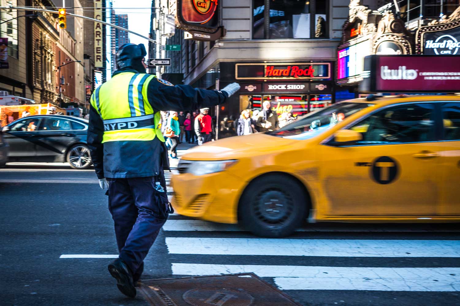 NYPD officer directs traffic as a yellow taxi crosses a bustling New York City intersection near Hard Rock Cafe.