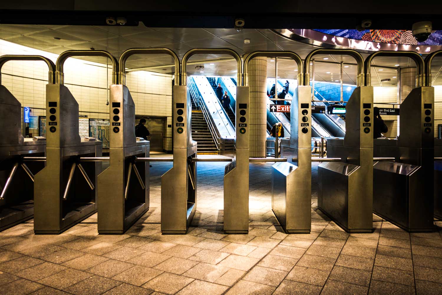 Subway turnstiles and escalators in a busy underground station, with passengers exiting and entering.
