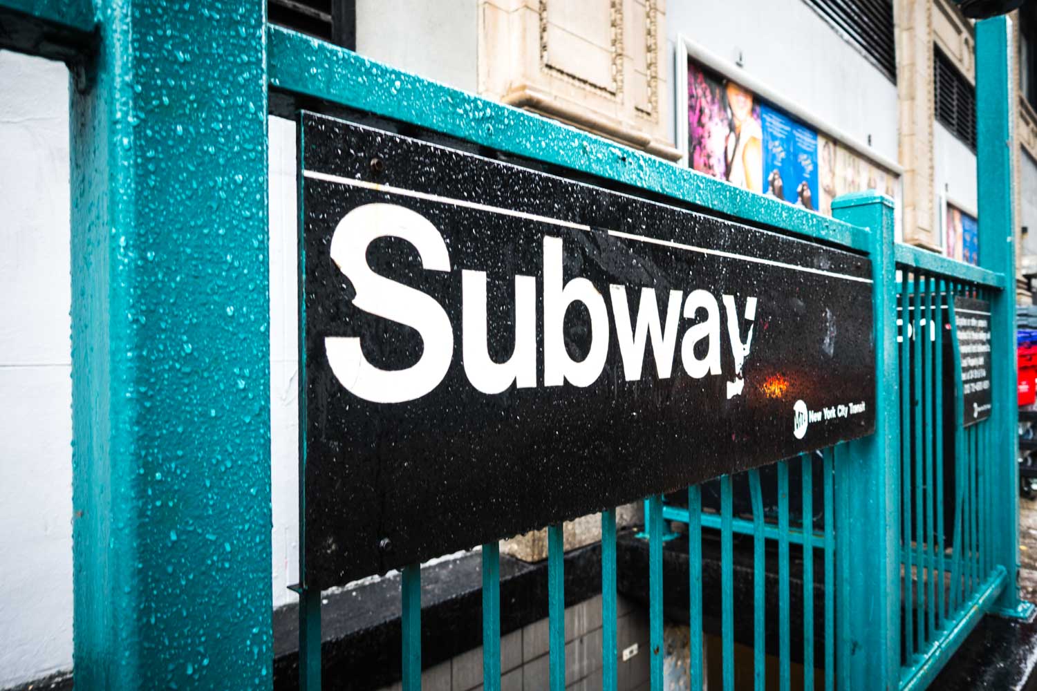 Rain-soaked New York City subway entrance with teal railings and bold signage.