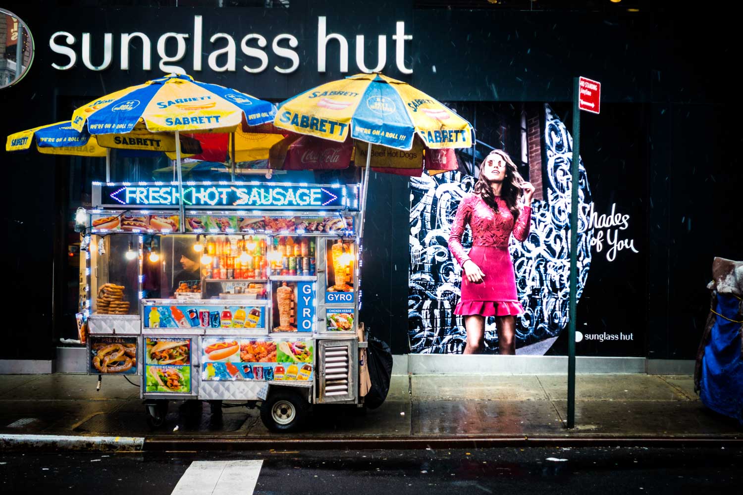 Food cart selling hot sausage and gyros under colorful umbrellas on a city street, with a fashionable ad in the background.