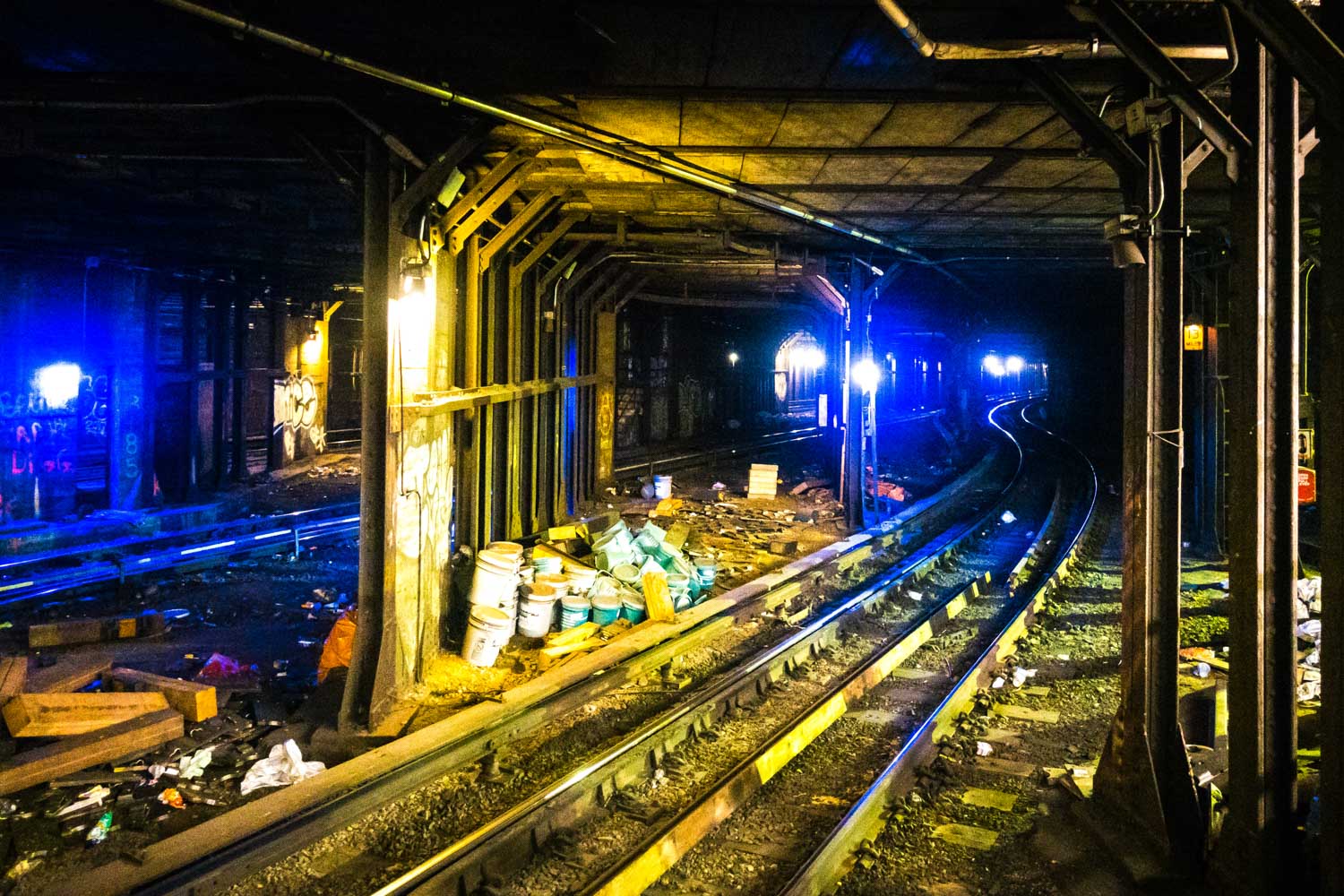 Subway tunnel with tracks, graffiti, and scattered trash, lit by yellow and blue lights for an urban underground atmosphere.