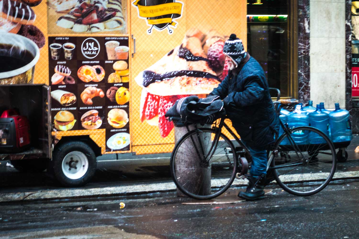 Person in winter gear stands with bike beside food cart displaying pastries and drinks on a wet city street.