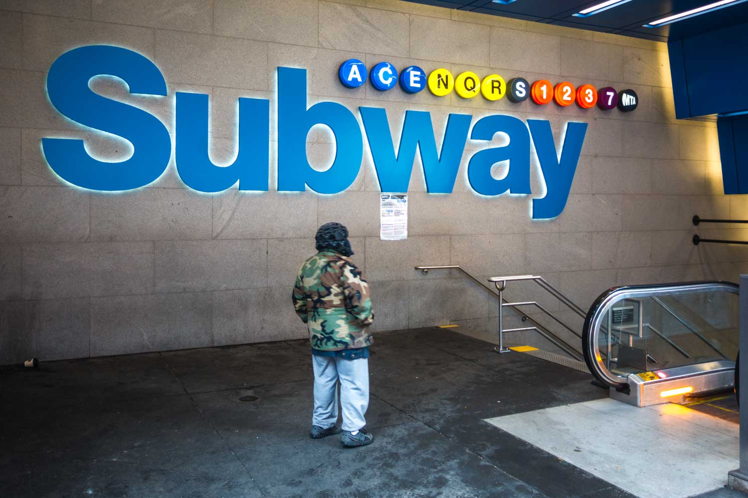 Person in camouflage jacket near large Subway sign with colorful line symbols, beside stairs and escalator entrance.