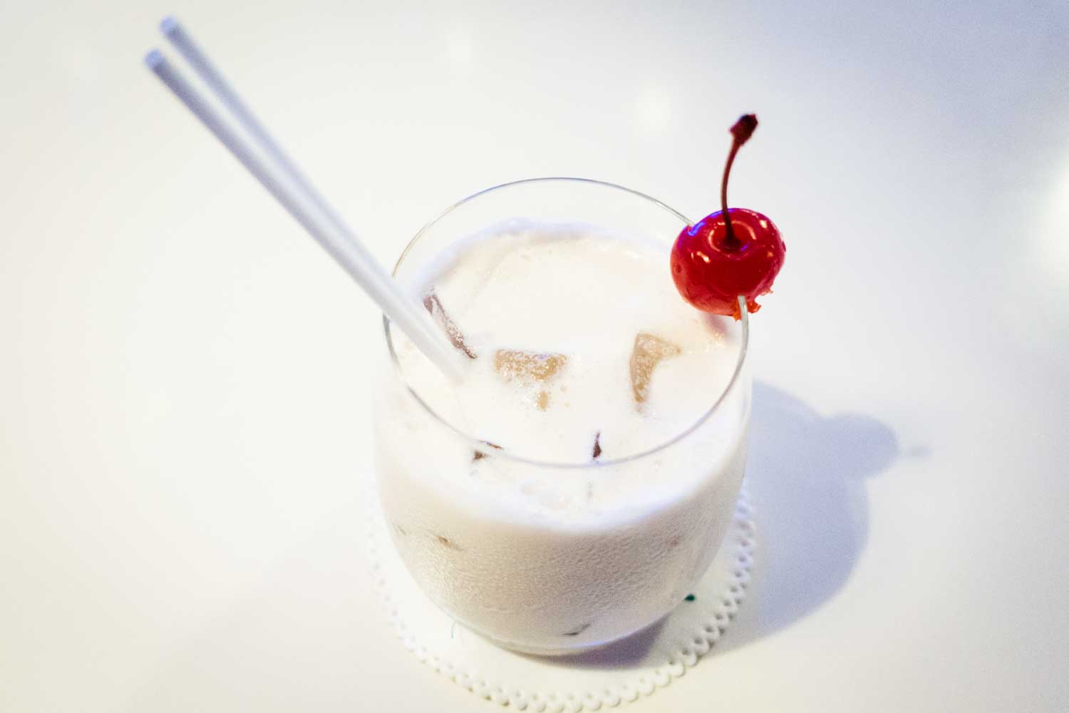 Creamy iced cocktail with a cherry garnish, served with two straws on a white table.