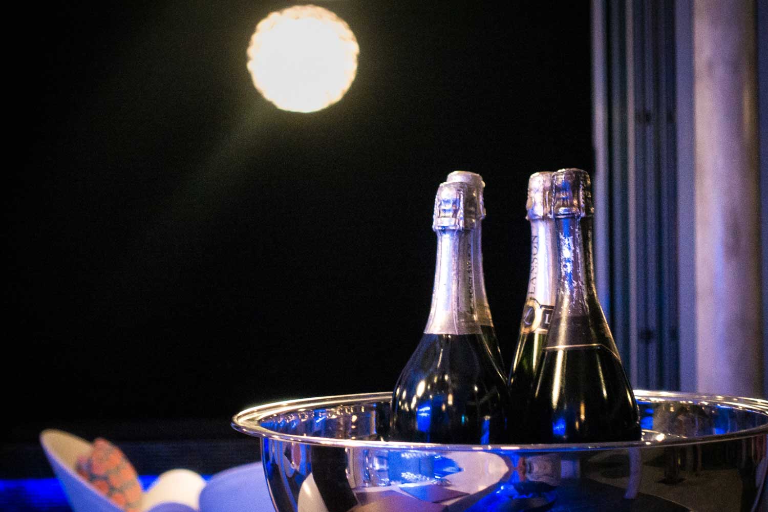 Two champagne bottles in an ice bucket under a glowing lamp at night, with modern seating in the background.