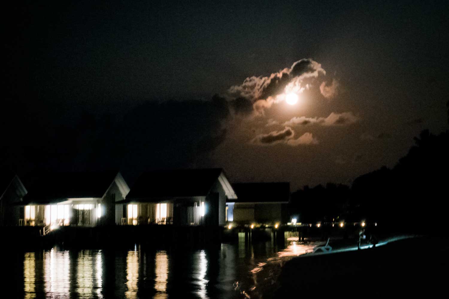Moonlit night over water villas, with glowing windows and reflections, under a cloudy sky.