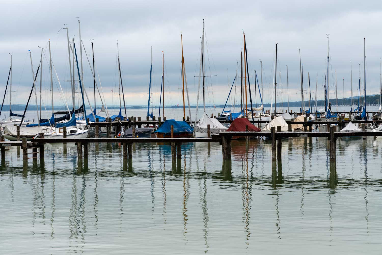 Sailboats with colorful covers docked at a serene marina, reflecting in calm water under a cloudy sky.