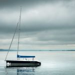 Sailboat with blue cover floating on calm lake under cloudy skies.