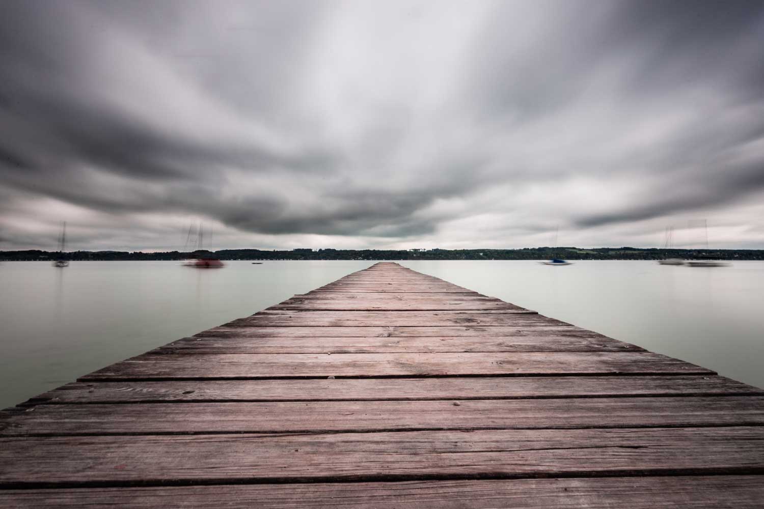 Wooden dock extending into calm lake under cloudy sky, with blurred boats in the distance.