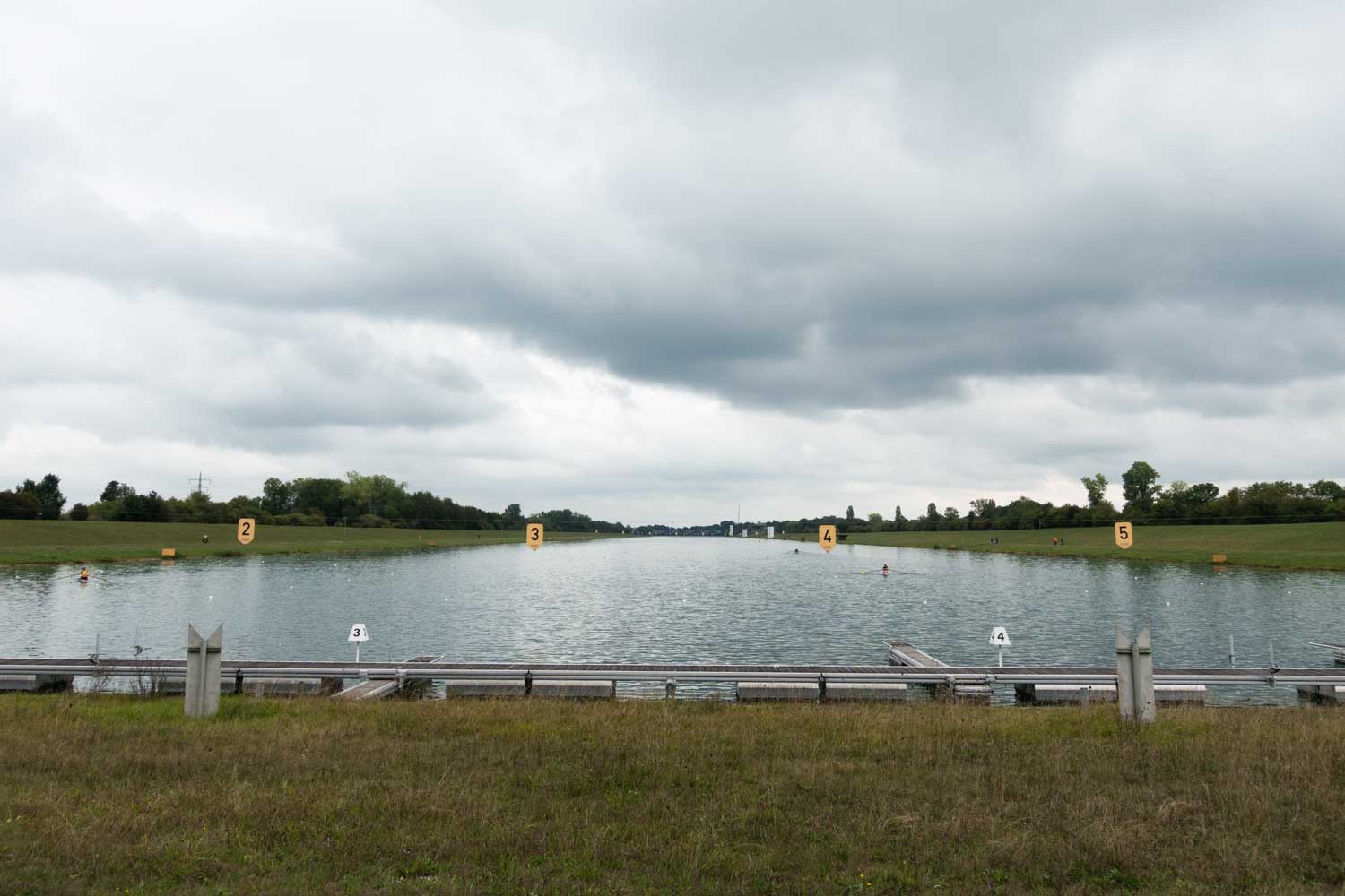 Wide view of a serene rowing canal under cloudy skies, with numbered lanes and grassy banks flanking the calm water.