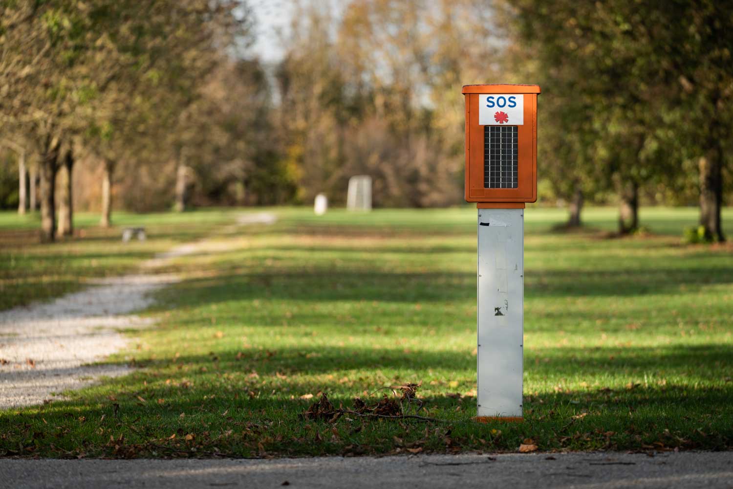 SOS emergency call box by a path in a grassy park, surrounded by trees in the background.