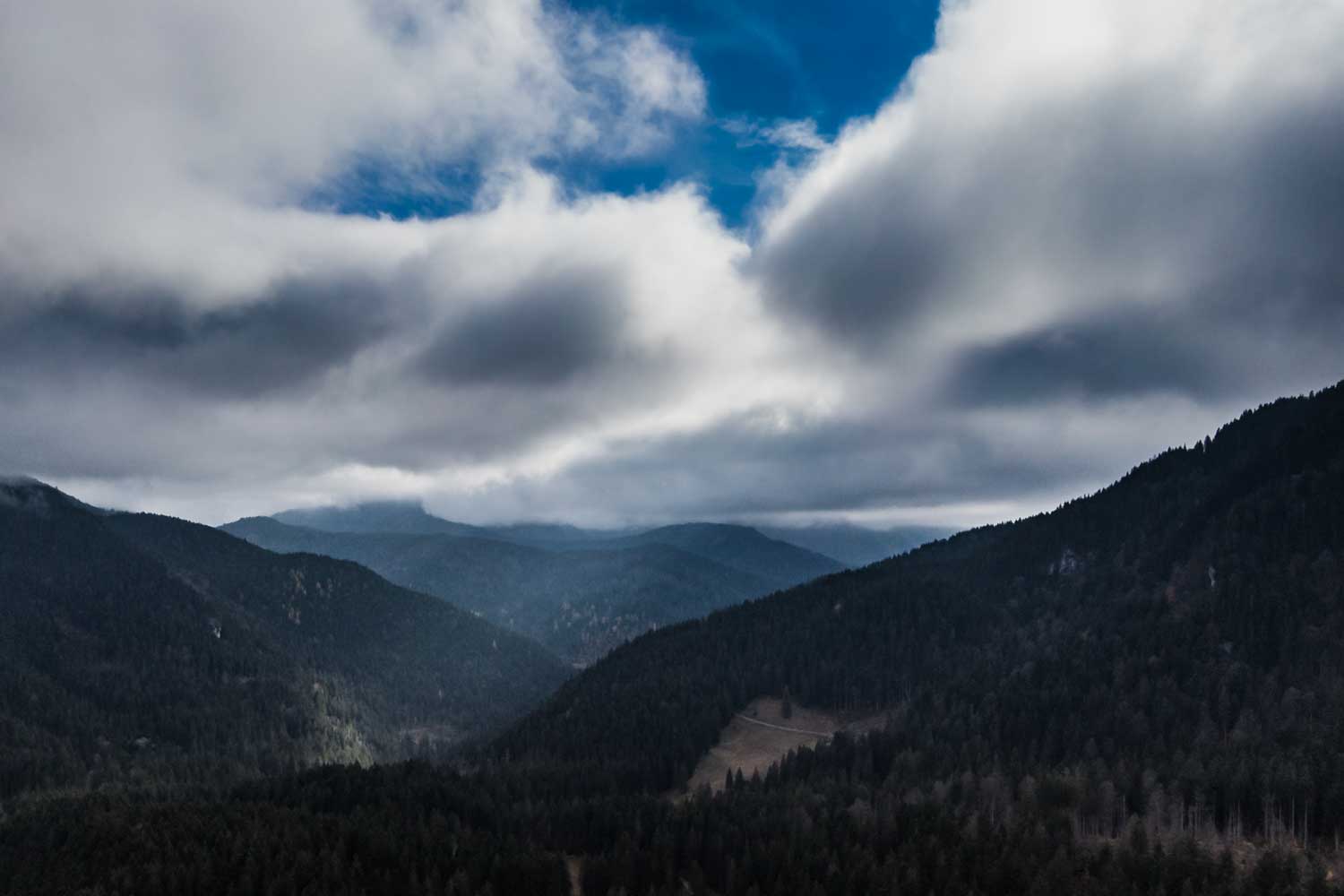 Mountain landscape with dense forests, clouds, and patches of blue sky, creating a serene and dramatic view.