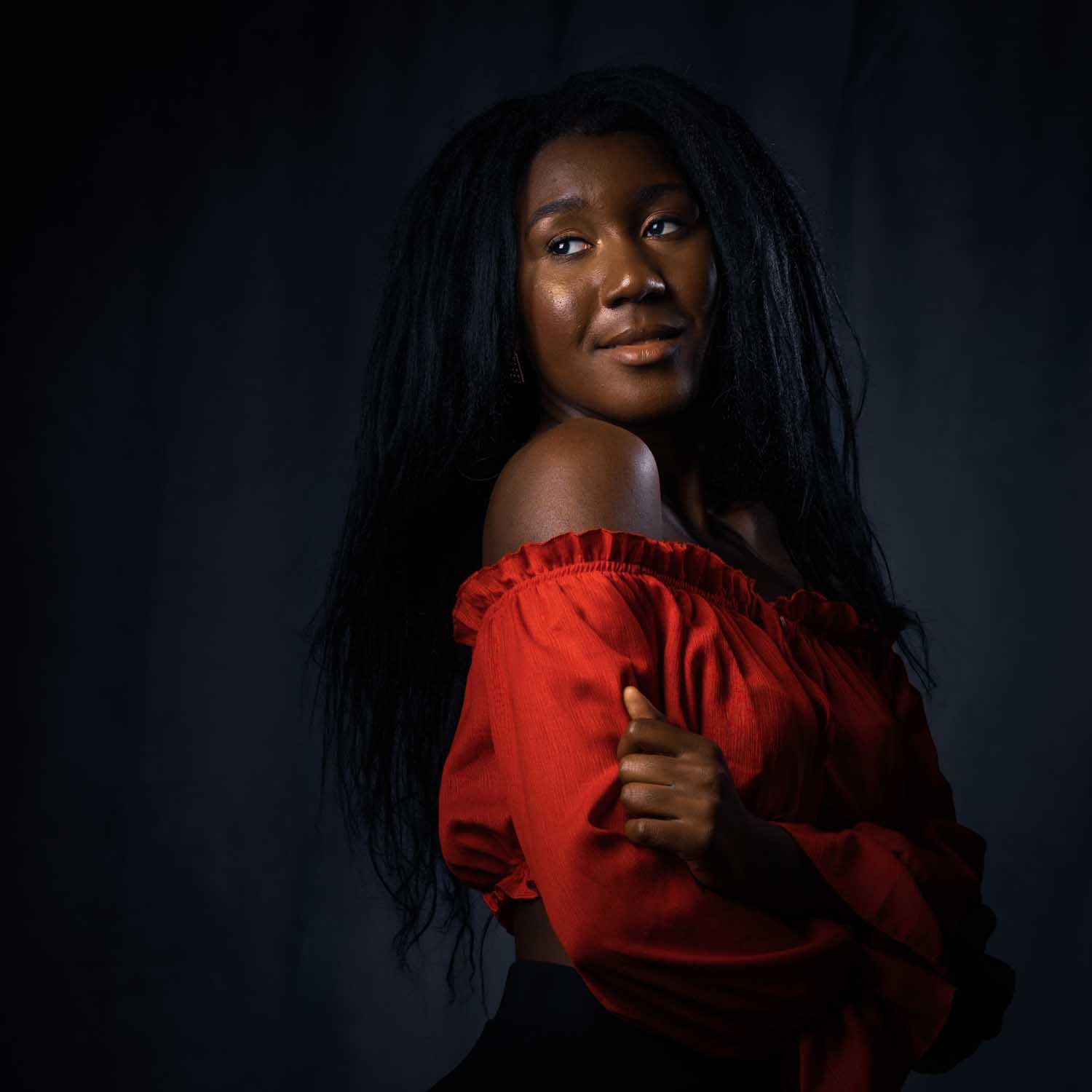 Woman in red off-shoulder top posing against a dark background, looking to the side.
