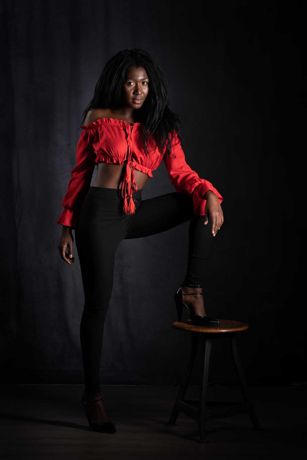 Woman in red crop top posing confidently on a stool against dark background.