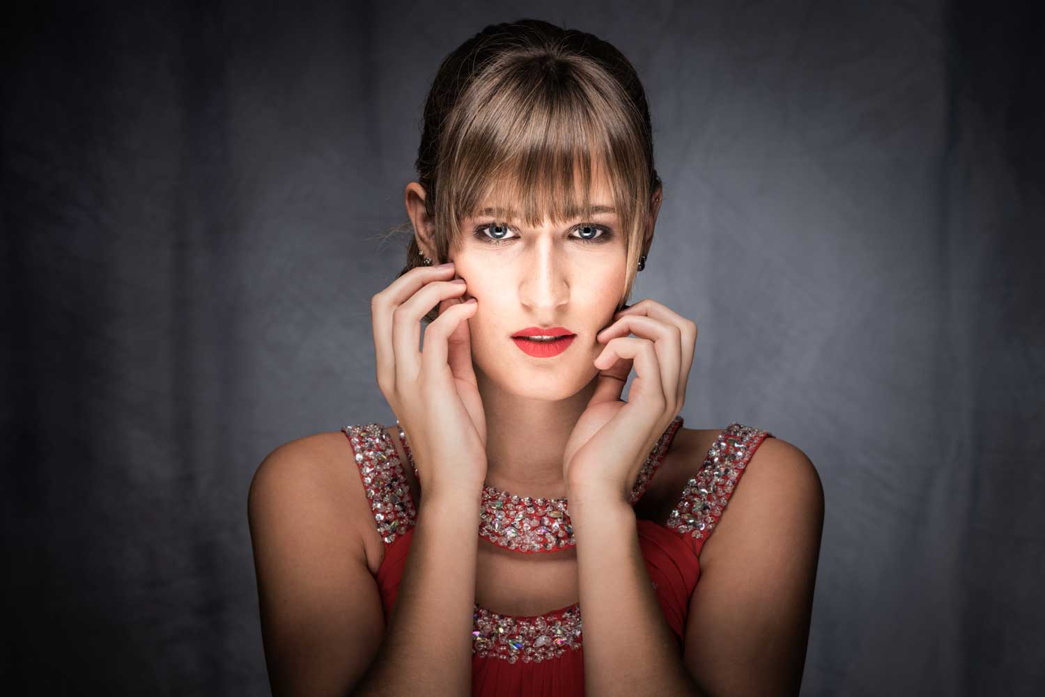 Glamorous woman with bangs and red lipstick, wearing a beaded red dress, posing in a dramatic studio setting.