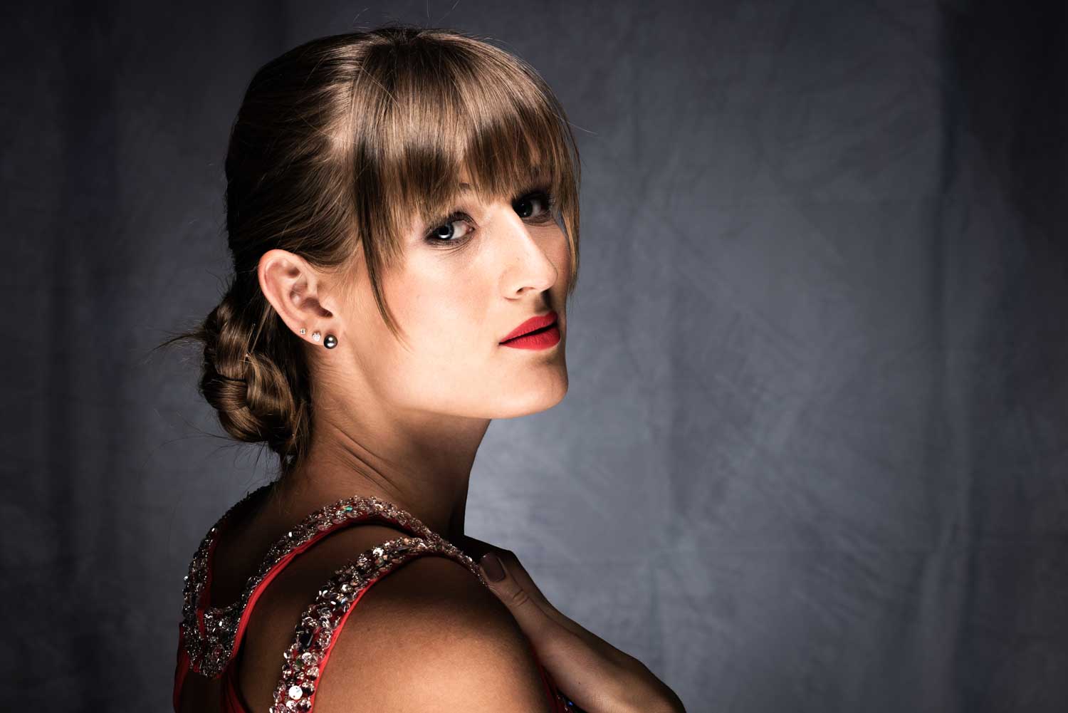 Woman with braided hair and fringe, wearing a jeweled dress against a gray background.