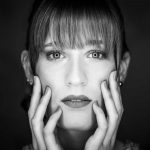 Black and white portrait of a woman with bangs, hands on her face, looking directly at the camera.