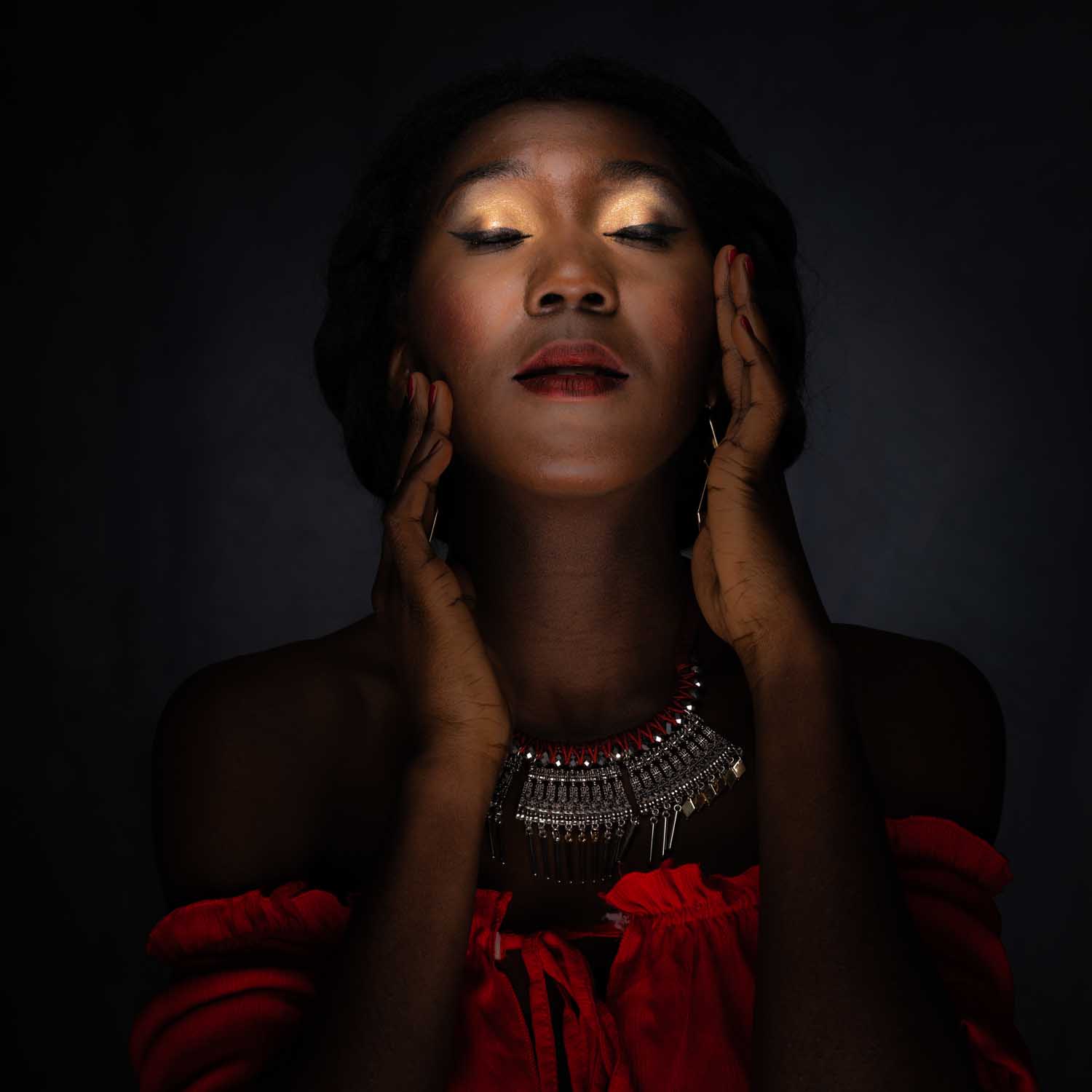 Woman in red dress and necklace with eyes closed, hands on face, under dramatic lighting.