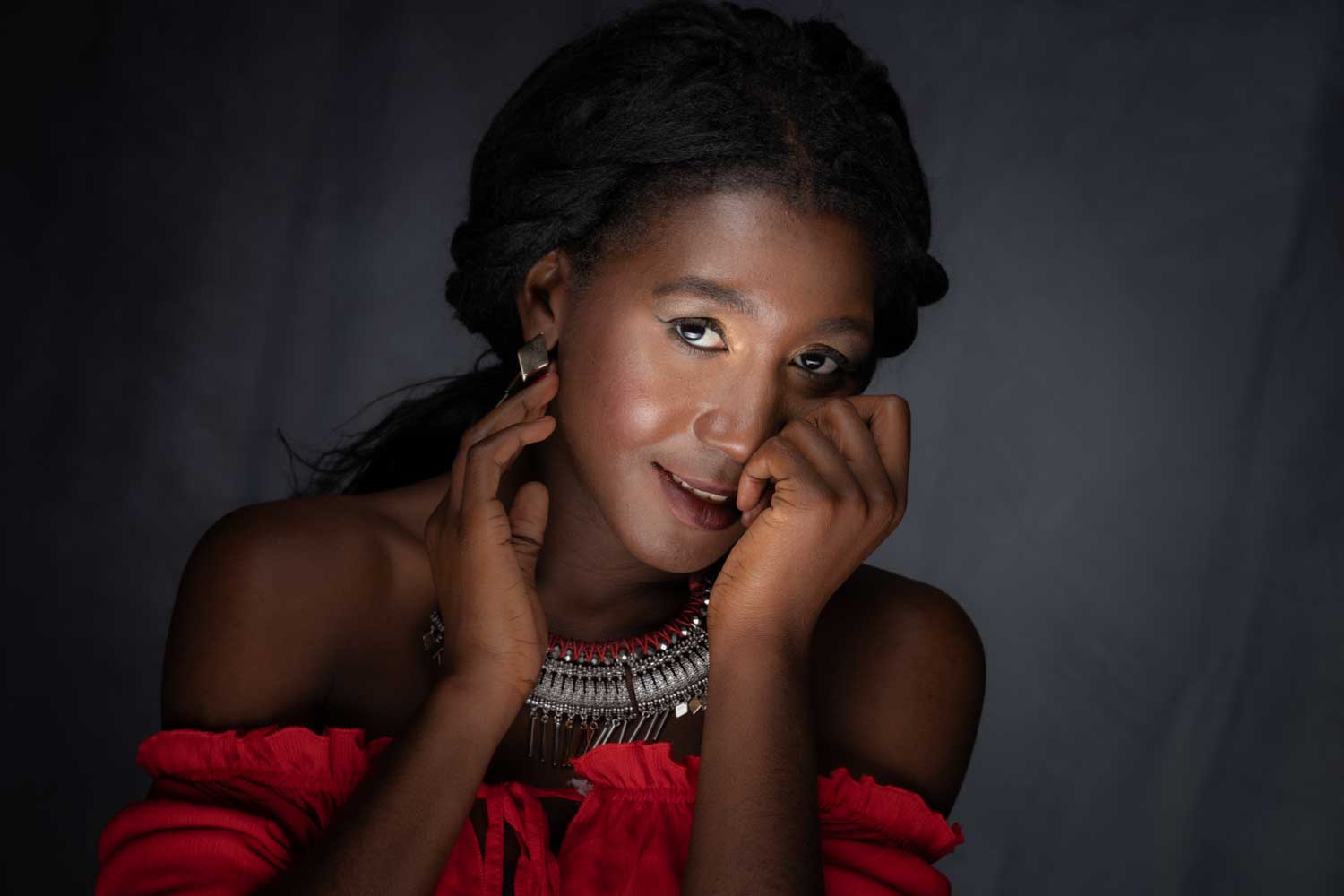 Woman in red off-shoulder top and statement necklace, smiling with hand on cheek against dark background.