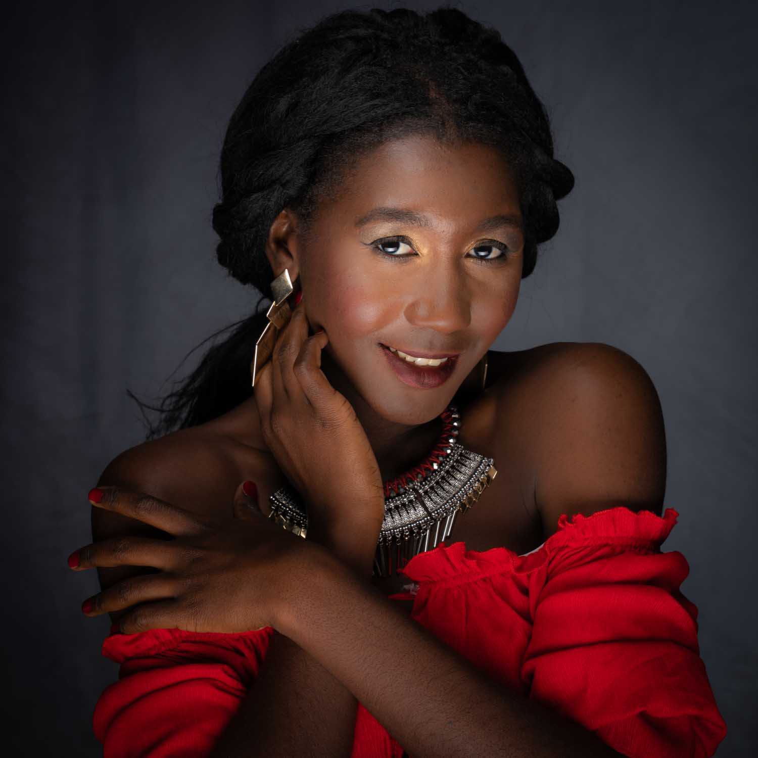 Smiling woman in red blouse with elaborate jewelry against dark background, hand touching face.