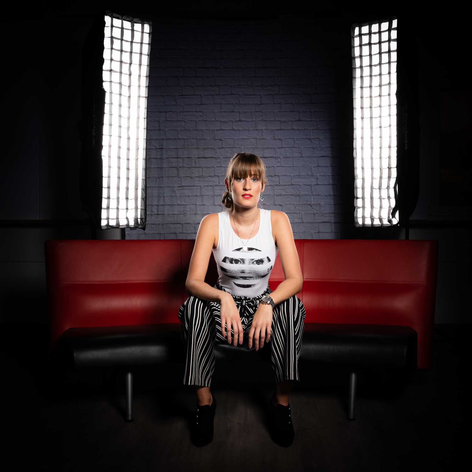 Woman in graphic tank top and striped pants sitting on a red couch, lit by studio lights against a brick backdrop.