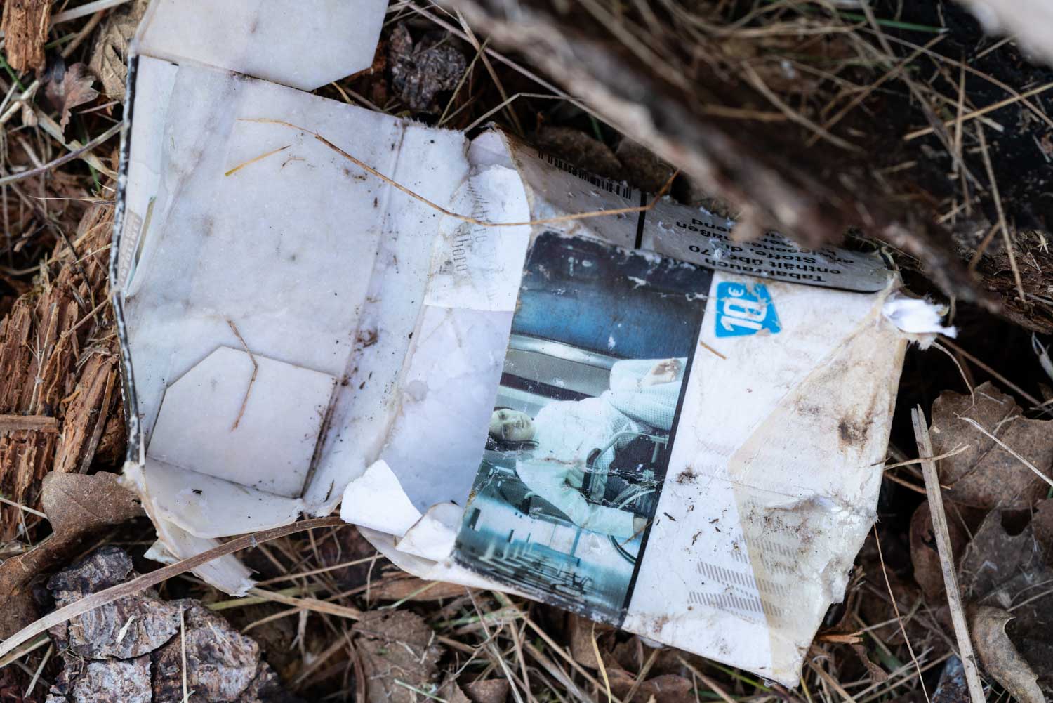 Discarded, worn magazine page with a photo of a person in white clothing, lying outdoors among dry leaves and branches.