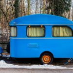 Blue vintage caravan with yellow wheels parked in snowy winter forest, featuring green curtains and surrounded by trees.