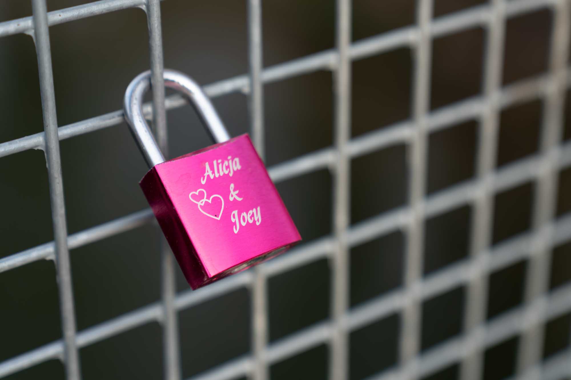 Pink love padlock engraved with hearts and names on a wire fence.