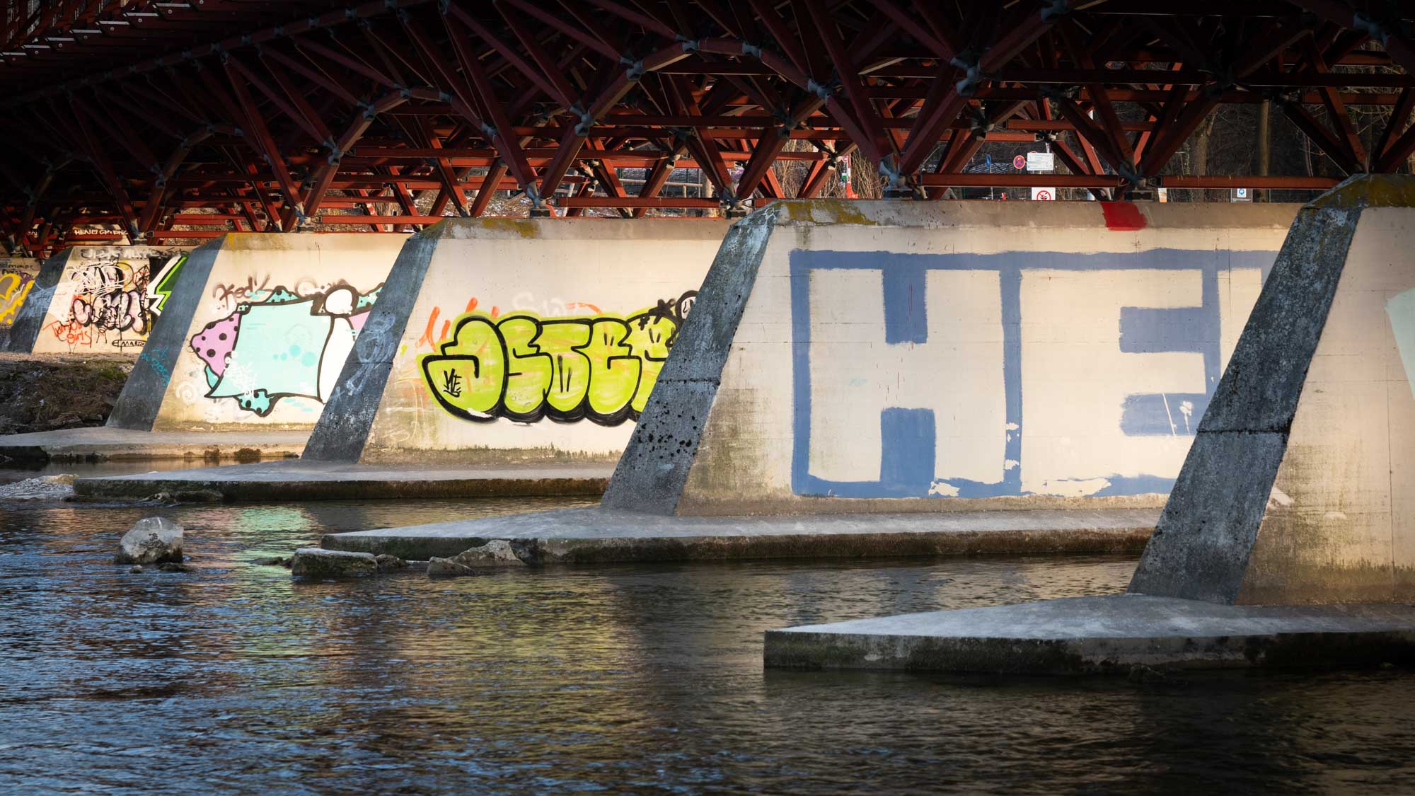Graffiti art on bridge pillars above water, featuring colorful designs and geometric shapes, under a metal framework.