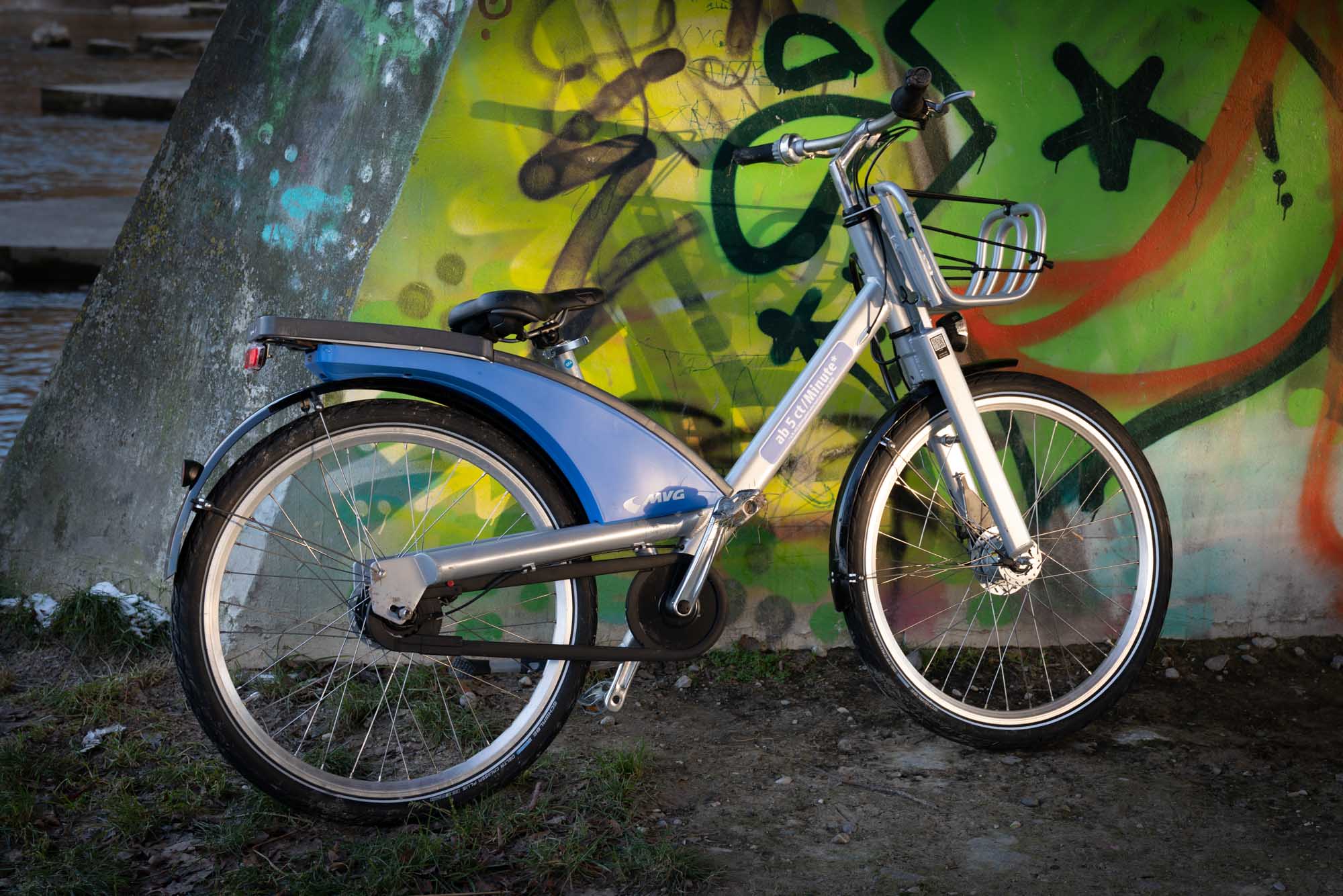 Blue rental bike parked by colorful graffiti wall near river.