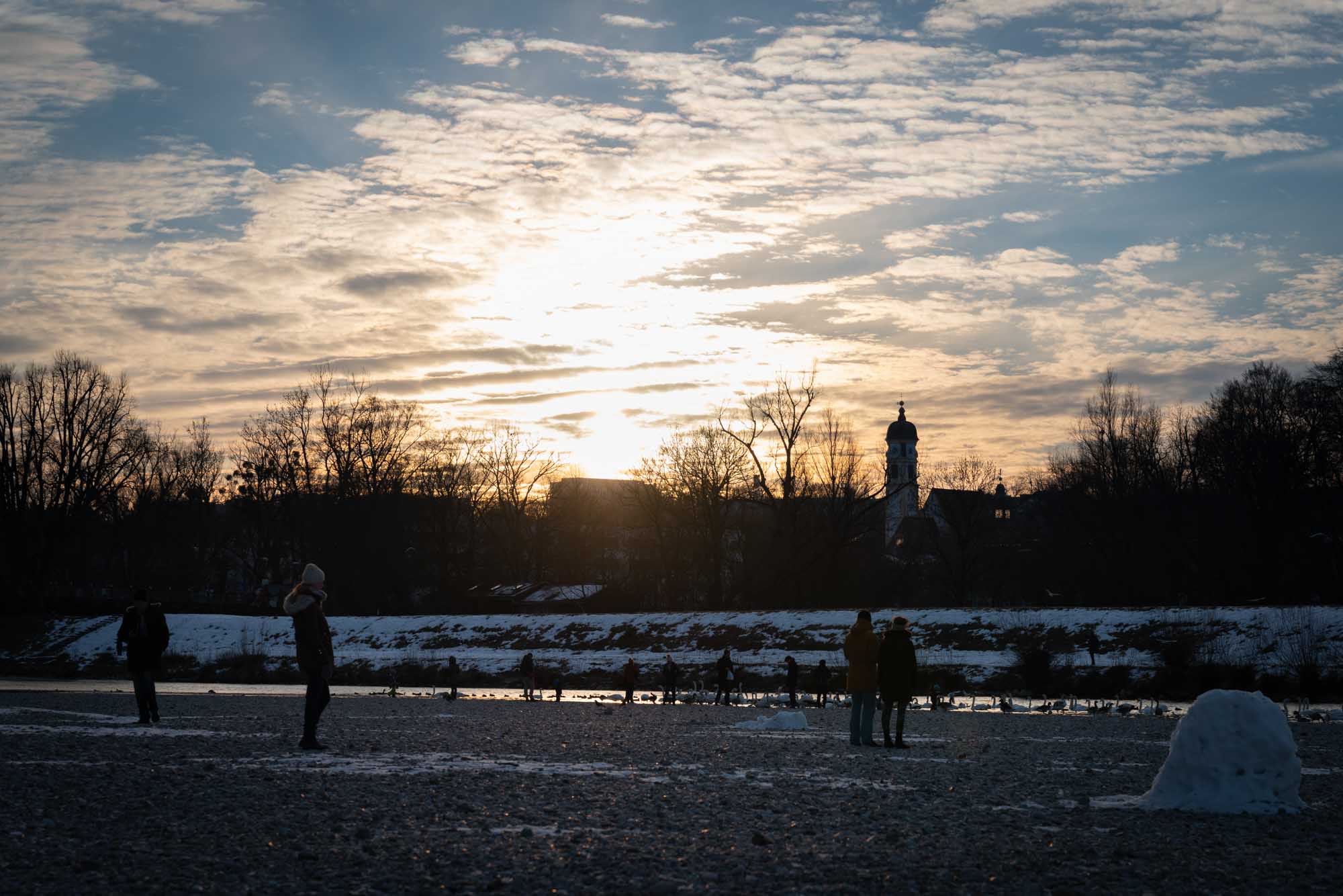 Winter sunset with silhouetted people on snowy ground, trees, and a distant church tower under a cloudy sky.