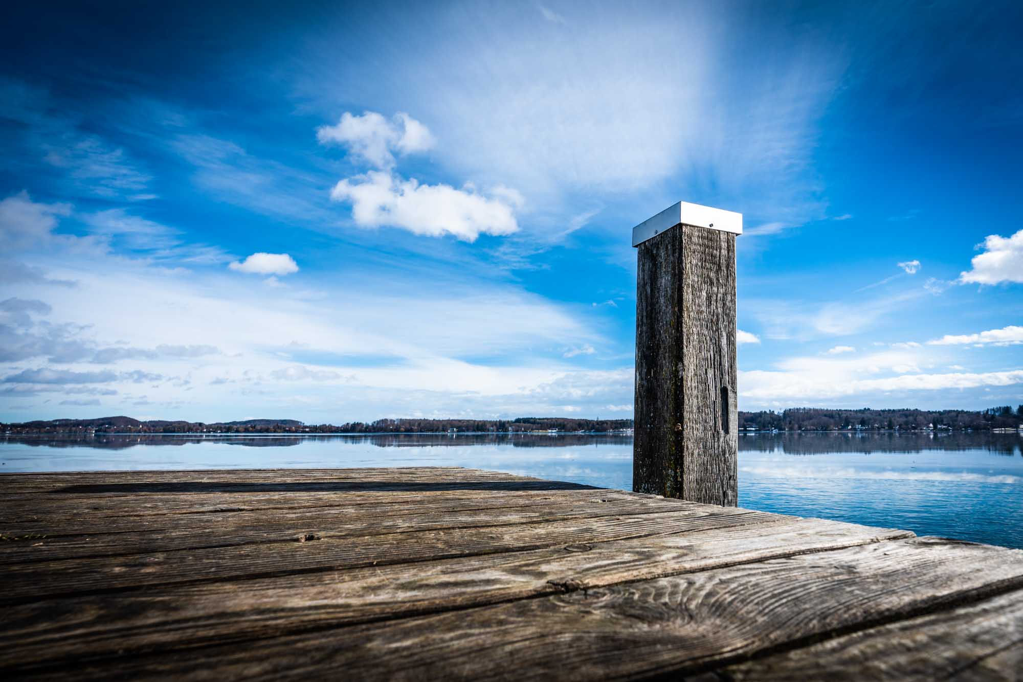 Tranquil lake view from a wooden dock against a vibrant blue sky with clouds.