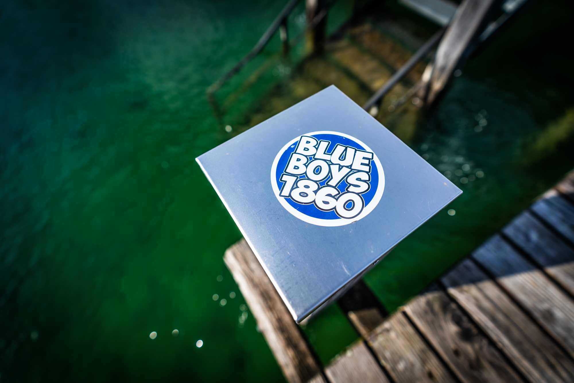 Sticker with Blue Boys 1860 logo on metallic surface by green lake water and wooden pier.